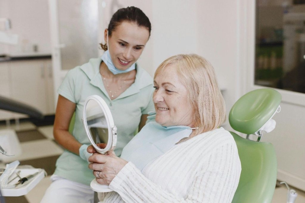 An older woman smiling while looking in a mirror after her dental appointment | What to Expect If You Haven’t Been to the Dentist in 10+ Years